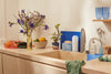 A sunny kitchen sink area with flowers and produce alongside blueland dish products