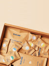 A cardboard box filled with several packets of Blueland products, including dishwasher detergent tabs and body wash. The packaging is brown with blue text and colorful circular labels. The background is a light beige color.
